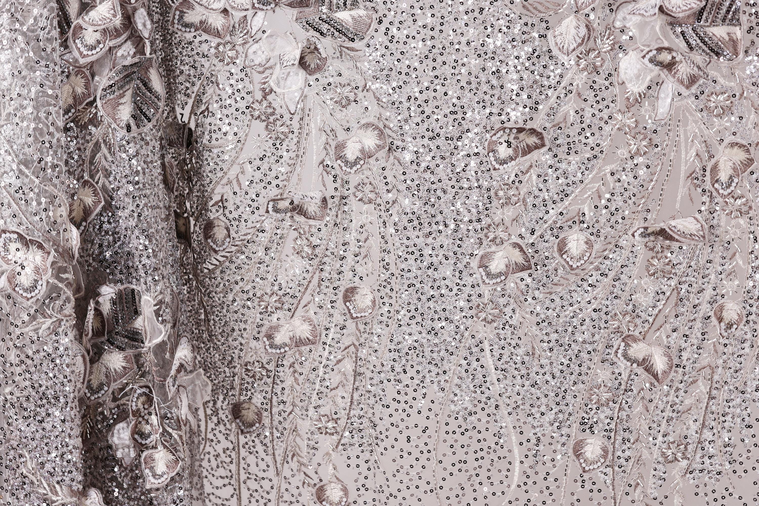 The image is a close-up of a fabric with a floral pattern, featuring a mix of small white and gray sequins and flowers.