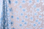 a close-up view of a fabric with a floral pattern. The fabric appears to be a light blue color, with white flowers and leaves scattered across it. The pattern is intricate and detailed, creating a visually appealing and textured surface.