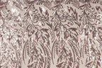 a close-up view of a fabric with a repeating pattern of brown and white sequin designs. The pattern appears to be a floral or leaf-like design, with the sequins arranged in a way that creates a sense of movement and texture. The fabric has a light brown or beige background, which contrasts with the dark brown and white colors of the sequins. The image does not contain any text or other discernible objects, and the focus is solely on the intricate and detailed pattern of the sequin design.