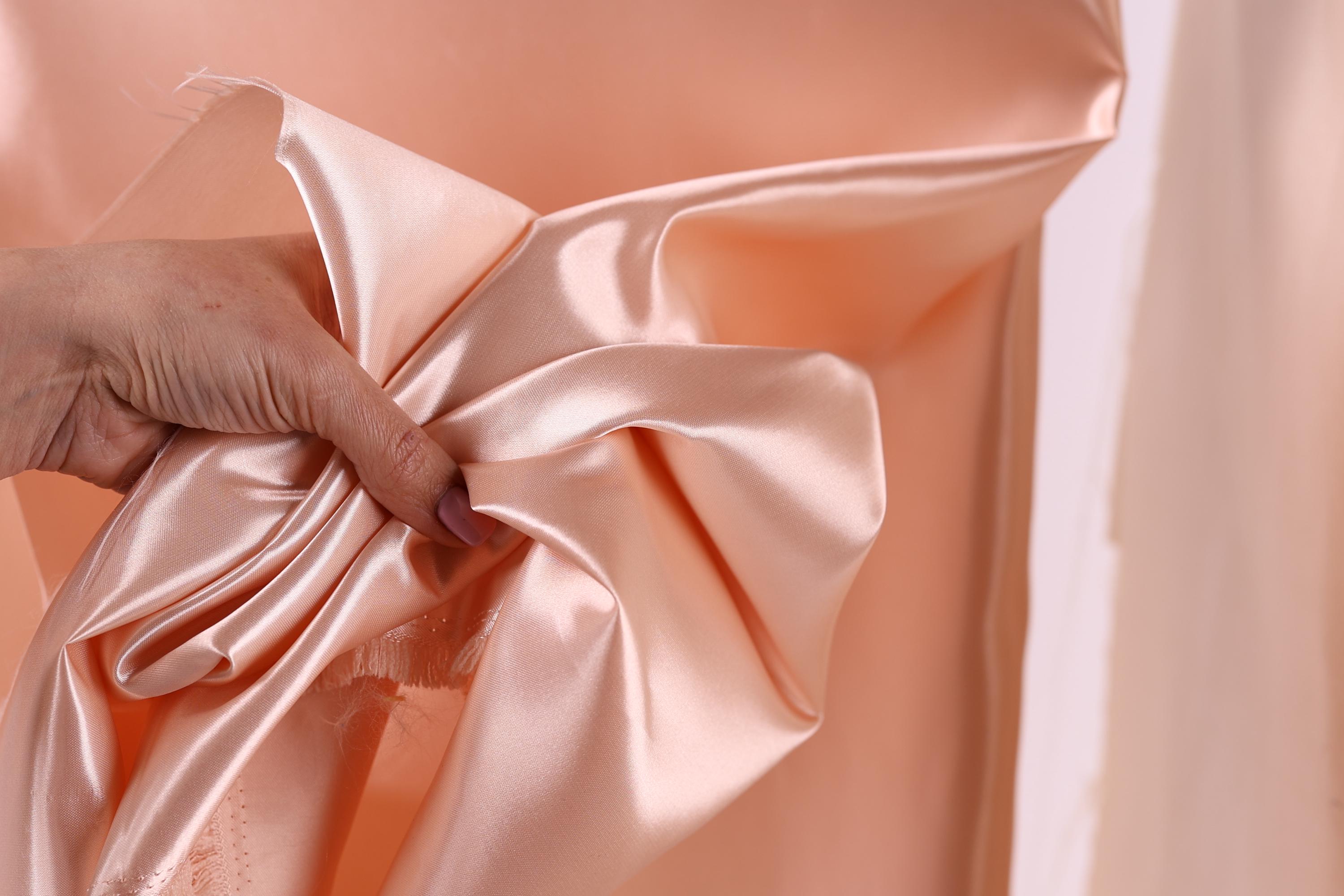 A hand is holding a piece of peach-colored satin fabric, which appears to be a bow or ribbon.