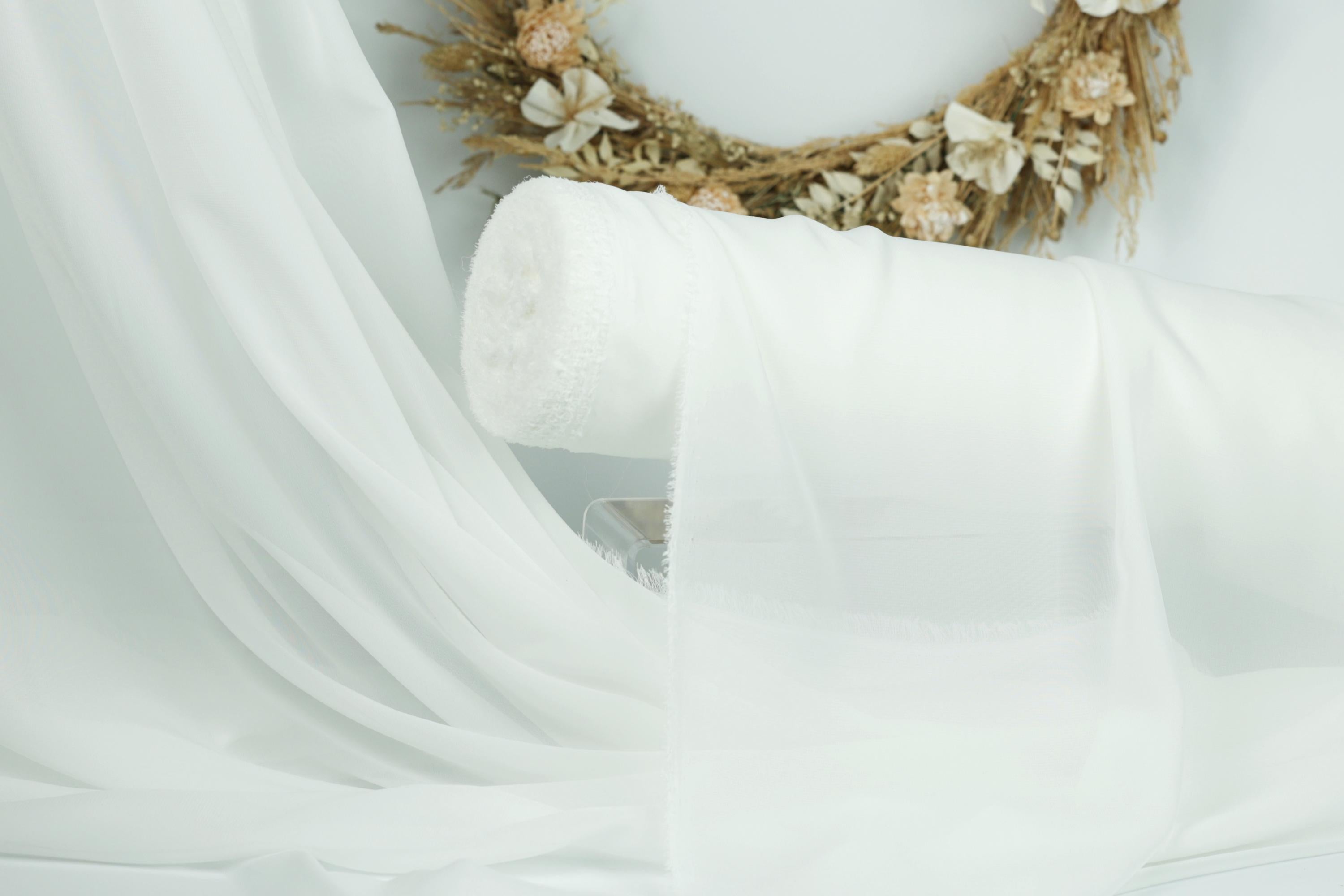 a white fabric, possibly a sheer or sheer curtain, draped over a white surface. In the background, there is a wreath made of dried flowers and branches, adding a decorative element to the scene.