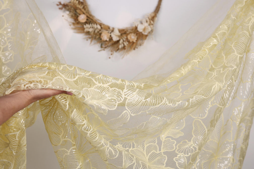 A hand is holding a yellow fabric with a floral pattern, which is draped over a wall. In the background, there is a wreath made of dried flowers and branches.