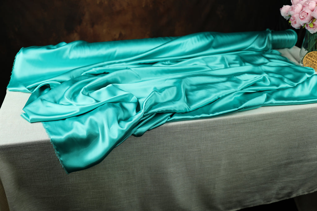 a roll of turquoise satin fabric on a table, with a vase of pink flowers visible in the background.