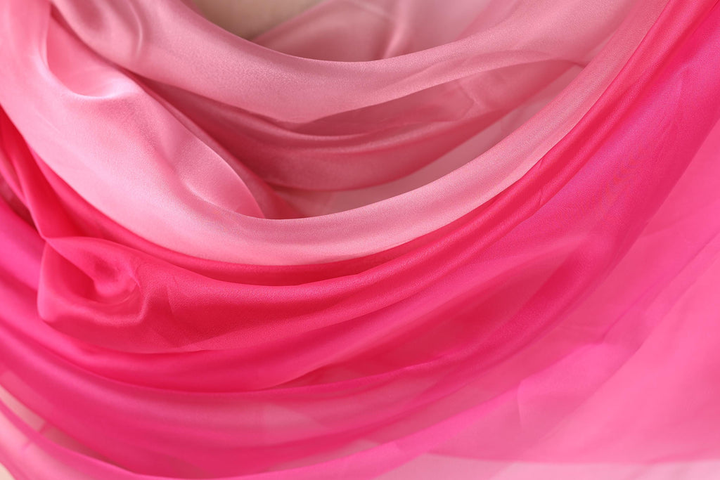 The image is a close-up of a soft, flowing fabric with a gradient of pink and white colors. The fabric appears to be made of silk or chiffon, and it has a smooth, shiny texture. The colors are vibrant and the fabric looks delicate and luxurious.