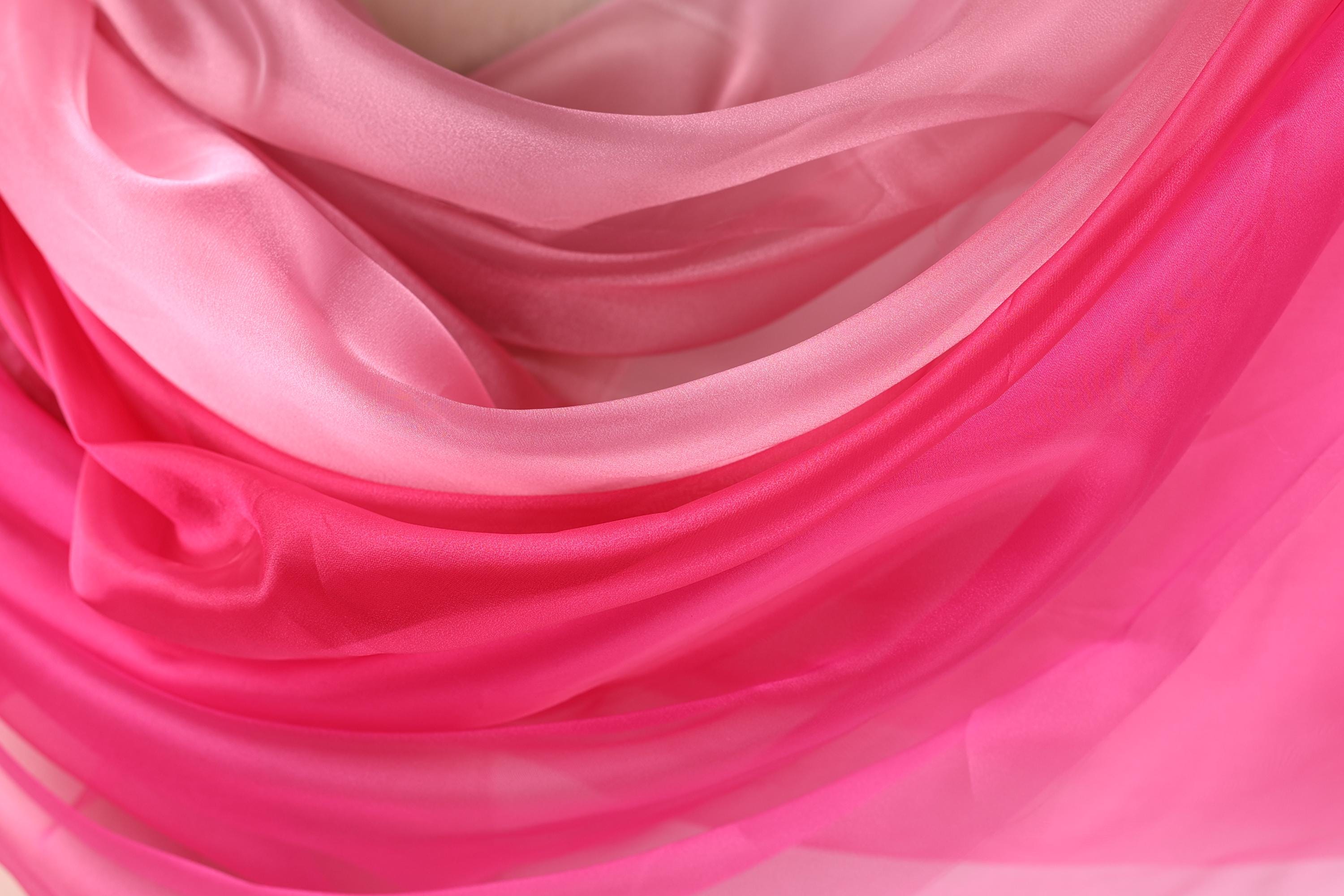 The image is a close-up of a soft, flowing fabric with a gradient of pink and white colors. The fabric appears to be made of silk or chiffon, and it has a smooth, shiny texture. The colors are vibrant and the fabric looks delicate and luxurious.