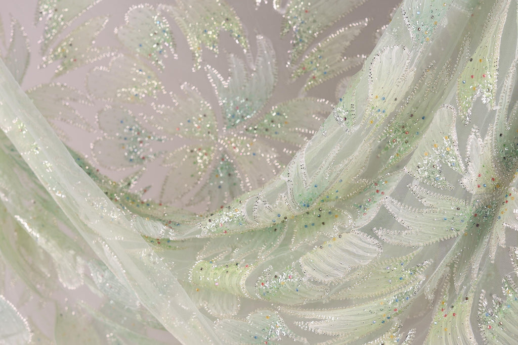 a close-up view of a delicate, light green fabric with a floral pattern. The fabric appears to be made of sheer material, with small, colorful beads or sequins scattered throughout the design. The overall composition of the image is visually appealing and showcases the intricate details of the fabric.