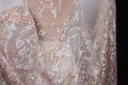 a close-up view of a delicate, sheer fabric with a floral pattern. The fabric appears to be made of lace or sheer material, with a pink and white color scheme. The fabric is partially obscured by a hand, adding a sense of depth and texture to the image.