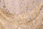 a close-up view of a delicate, intricately embroidered fabric with a floral pattern. The fabric appears to be made of sheer material, giving it a soft and delicate appearance. The embroidery is detailed and colorful, with various shades of pink, yellow, and green creating a vibrant and eye-catching design. The fabric is spread out, showcasing the full extent of the embroidered pattern and its intricate design.