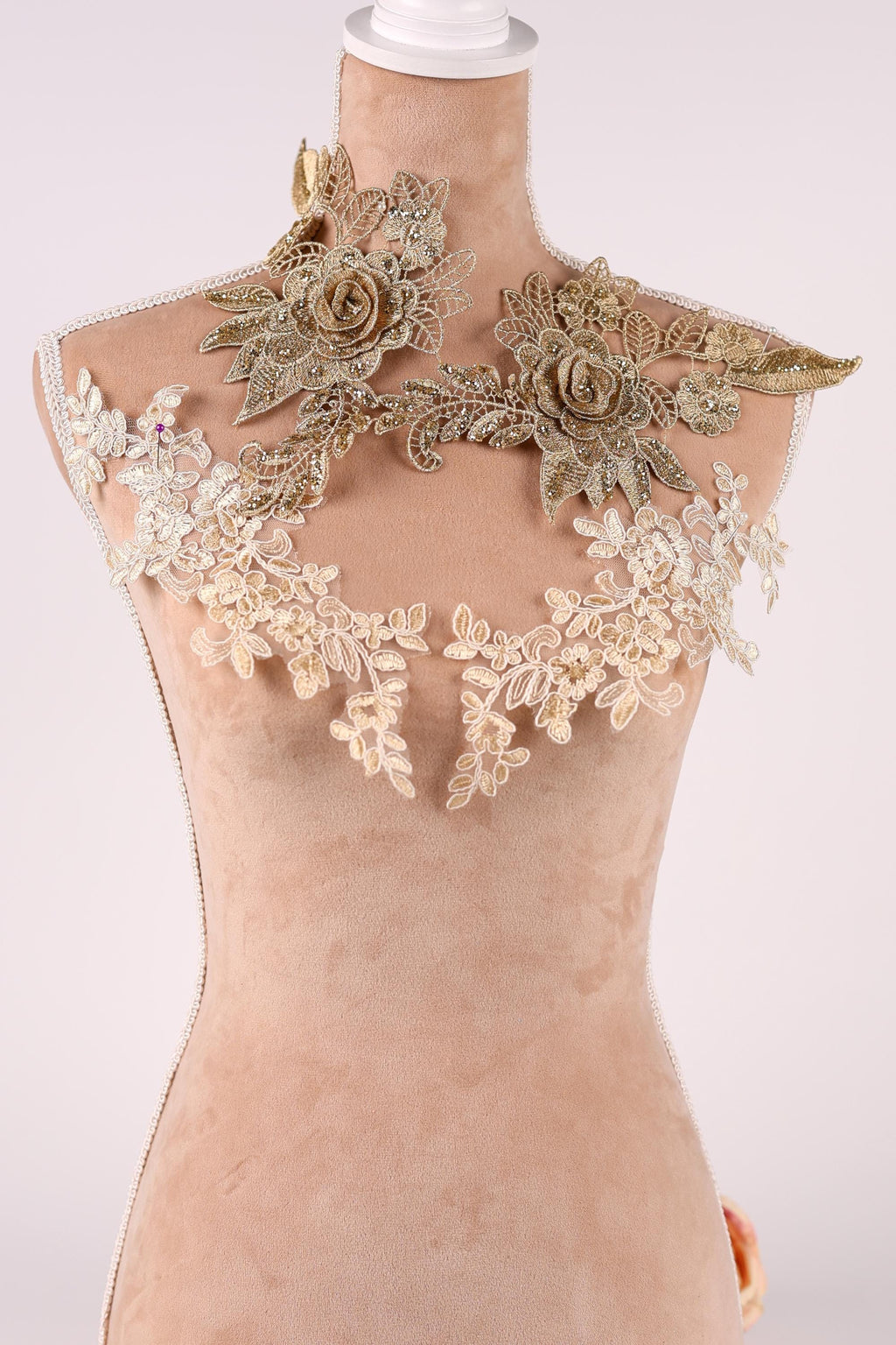 A mannequin wearing a beige dress with intricate gold and white lace details, including floral and leaf-like patterns.
