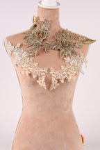 A mannequin wearing a beige dress with intricate gold and white lace details, including floral and leaf-like patterns.