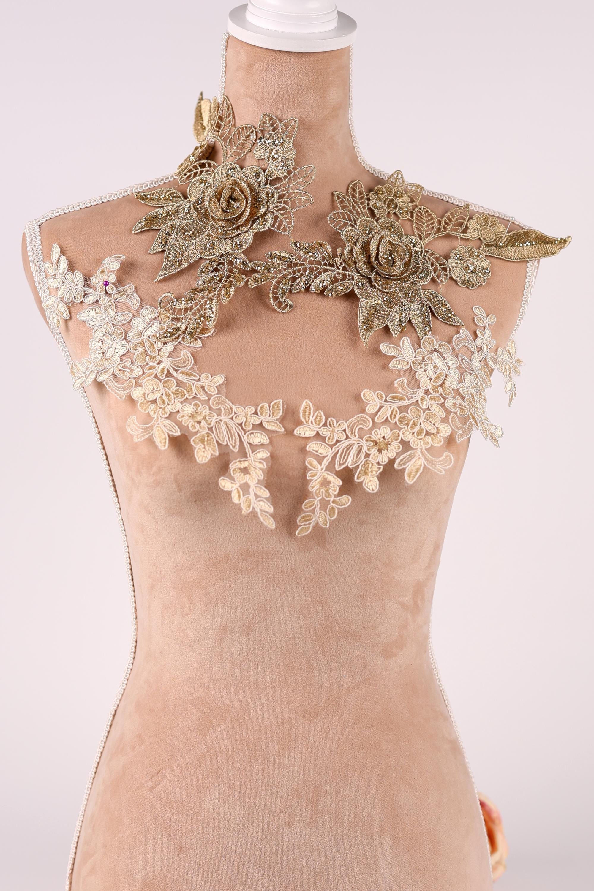 A mannequin wearing a beige dress with intricate gold and white lace details, including floral and leaf-like patterns.