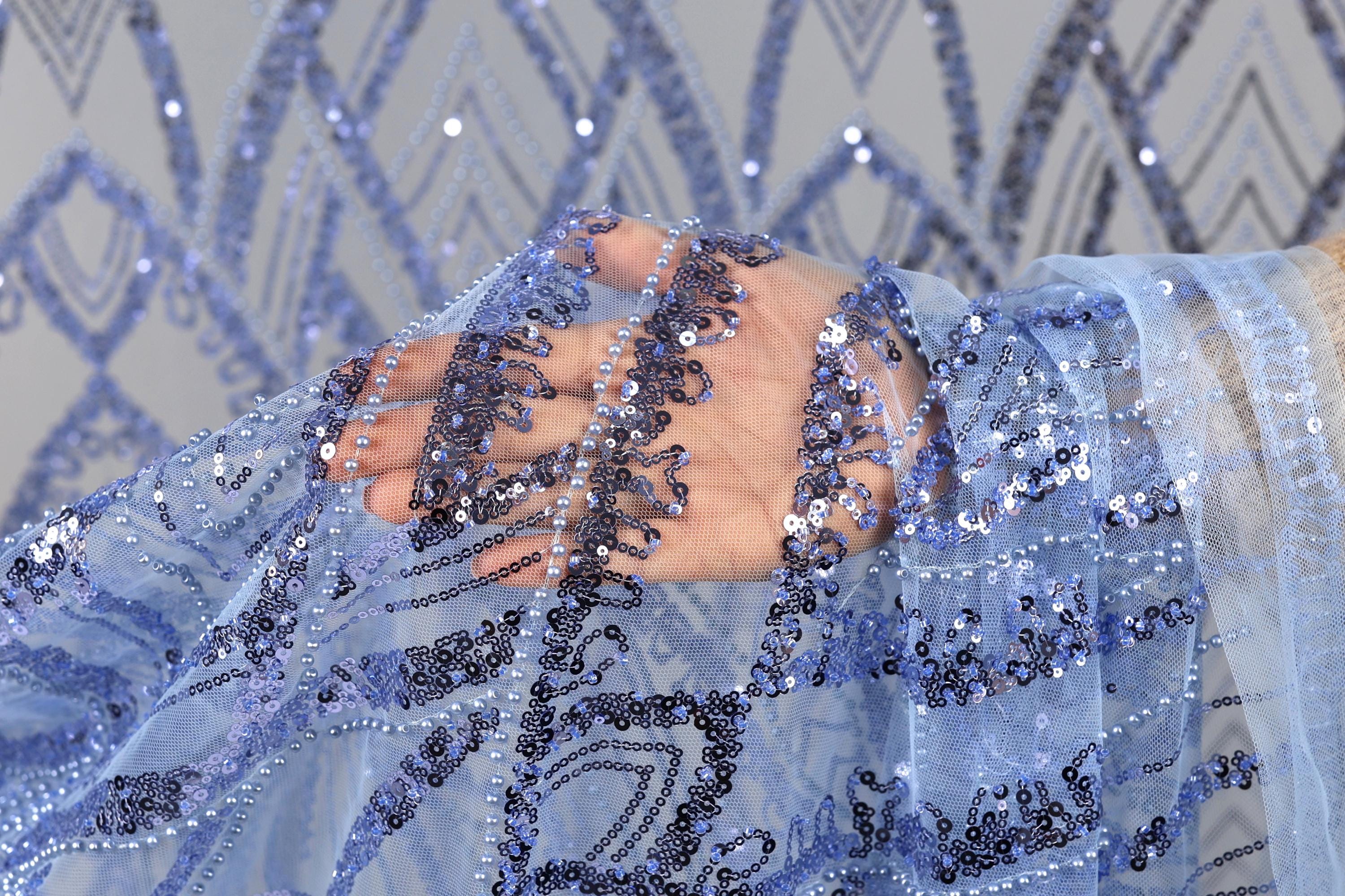 A hand is holding a piece of blue fabric with sequin details, against a background of similar blue and white sequin patterns.