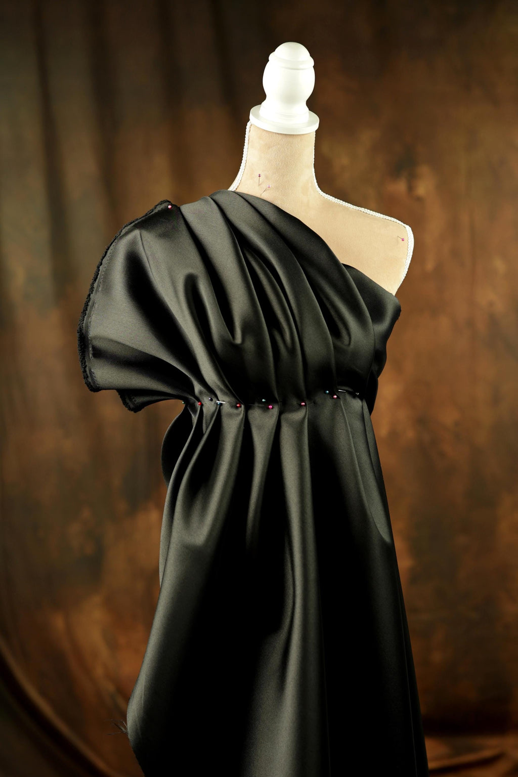 A black one-shoulder dress displayed on a mannequin against a brown background.