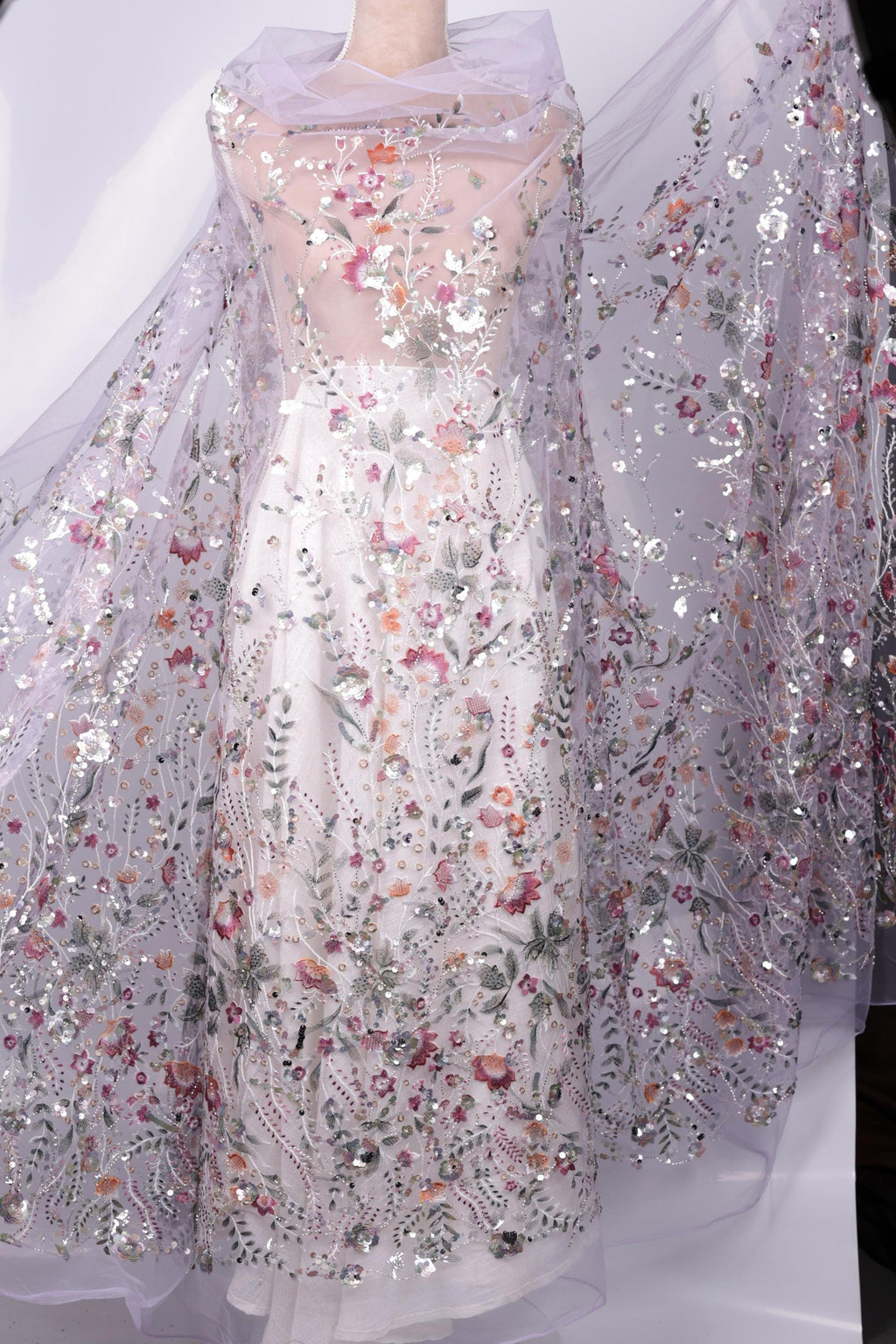 a long, white dress with a sheer, floral-patterned fabric draped over it. The dress has a sheer, transparent overlay that adds an elegant and delicate touch to the overall design.