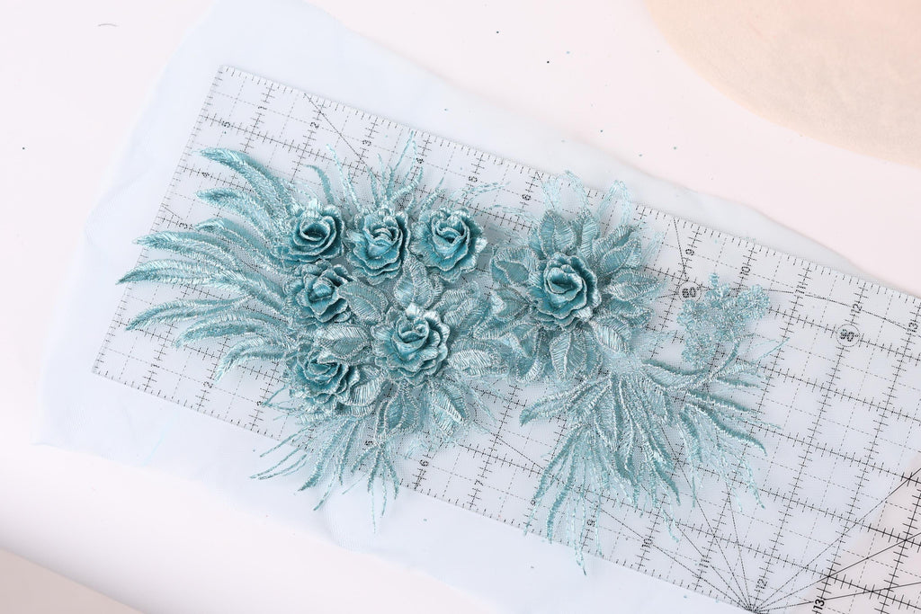 a close-up view of a piece of fabric with delicate blue floral designs, including multiple rose-shaped flowers and leaf-like elements. The fabric appears to be a light blue color, and the design elements are arranged on a white surface, possibly a cutting mat or ruler, which is placed on a white background.