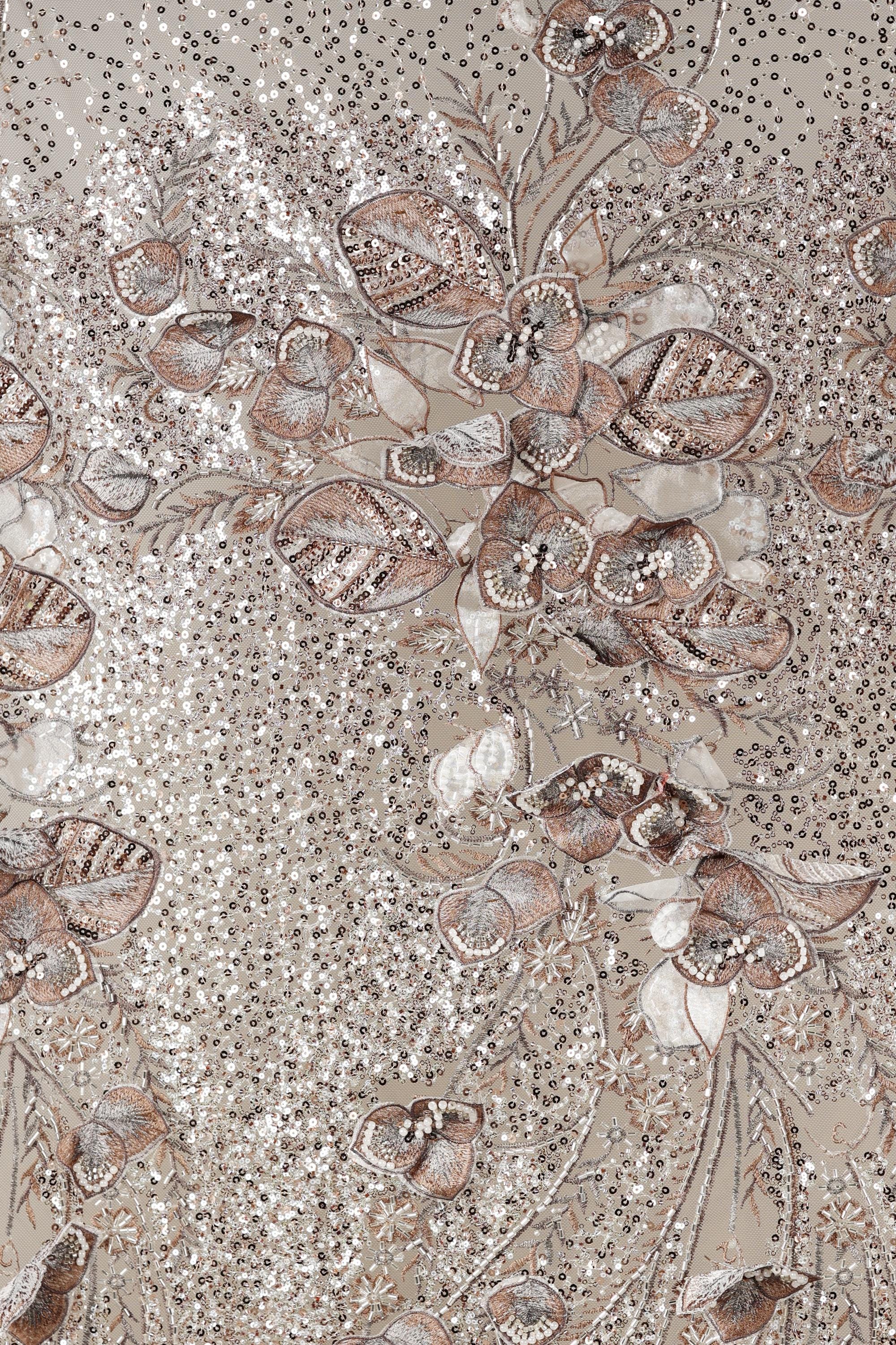 The image is a close-up of a fabric with a floral pattern, featuring a variety of flowers and leaves in shades of brown and beige. The pattern is intricate and detailed, with the flowers and leaves appearing to be made of sequins or other embellishments. The overall composition of the image is visually appealing and showcases the craftsmanship of the fabric design.