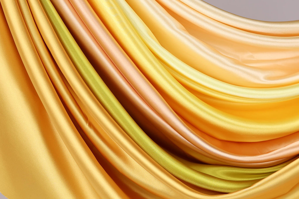 a close-up view of a vibrant, flowing fabric with a gradient of yellow and orange colors. The fabric appears to be made of silk or satin, and it is draped in a way that creates a sense of movement and dynamism. The colors are rich and warm, with the yellow and orange hues ranging from light to dark, creating a visually striking contrast. The fabric's texture and folds add depth and interest to the image, making it an eye-catching and artistic composition.