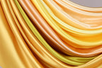 a close-up view of a vibrant, flowing fabric with a gradient of yellow and orange colors. The fabric appears to be made of silk or satin, and it is draped in a way that creates a sense of movement and dynamism. The colors are rich and warm, with the yellow and orange hues ranging from light to dark, creating a visually striking contrast. The fabric's texture and folds add depth and interest to the image, making it an eye-catching and artistic composition.