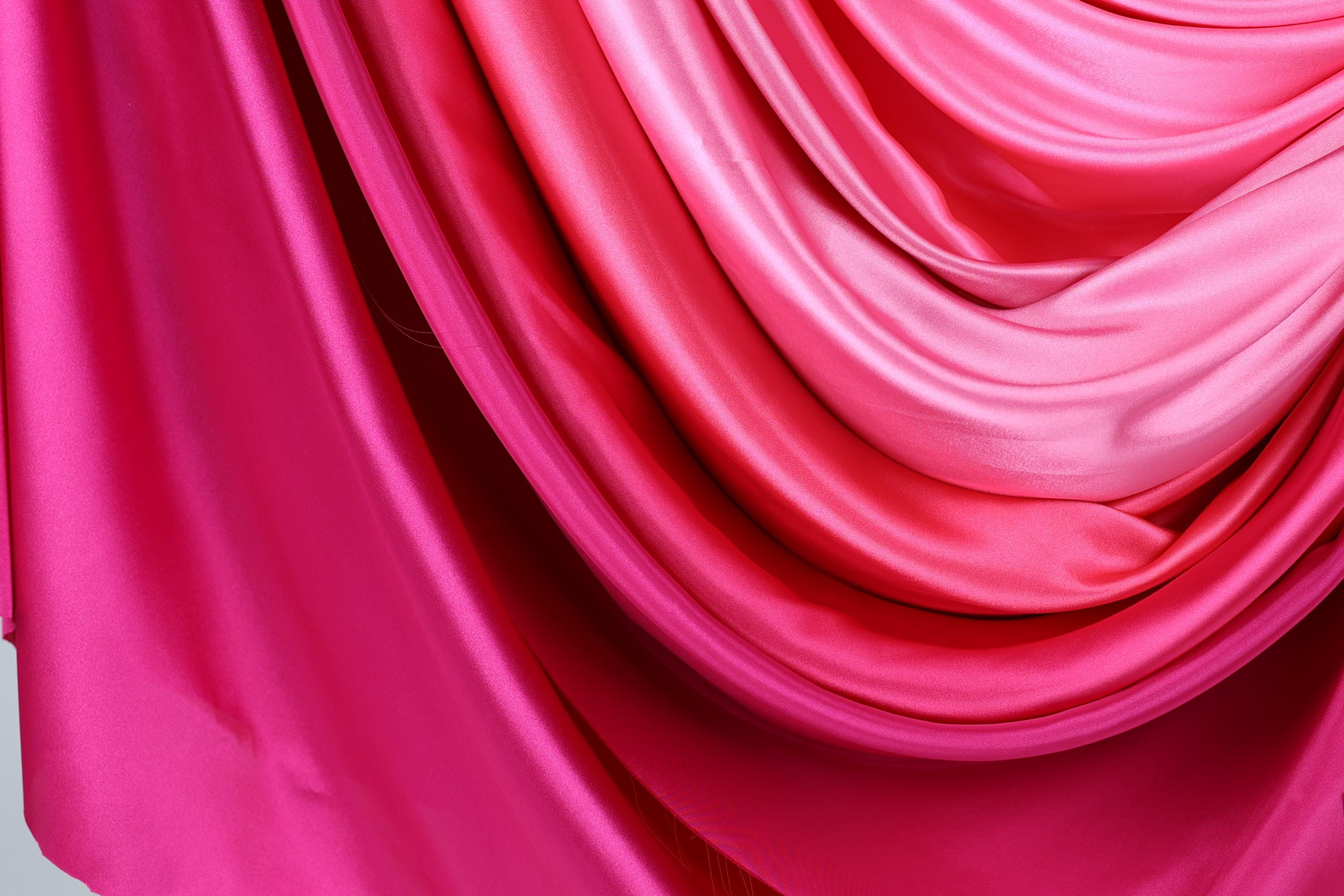 a close-up view of a vibrant, flowing fabric with a gradient of pink and red colors. The fabric appears to be made of silk or satin, and it is draped in a way that creates a sense of movement and elegance. The colors are rich and intense, with the pink and red hues dominating the fabric, while the lighter pink and red tones are more subtle, adding depth and dimension to the overall appearance.