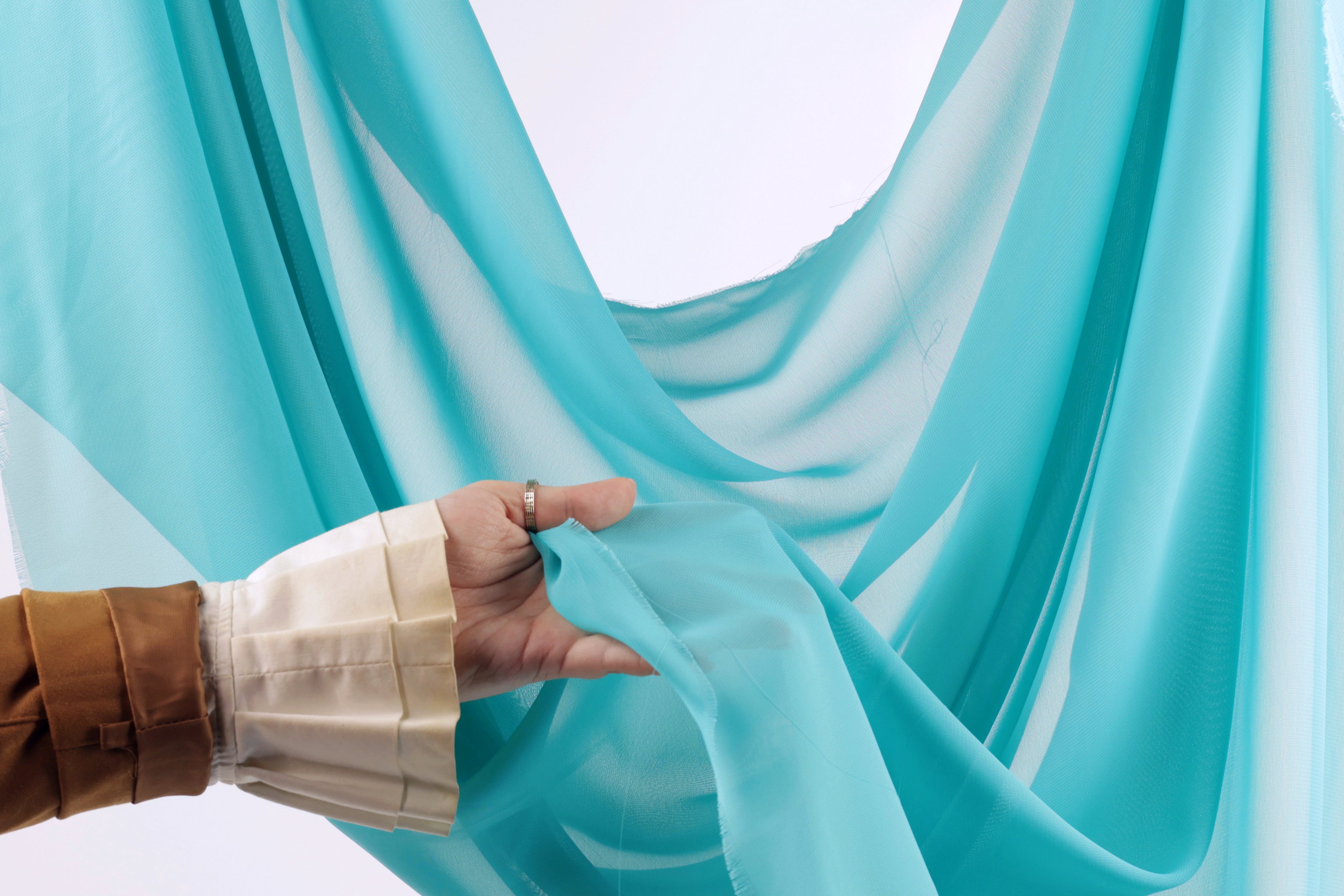 A hand is holding a piece of blue fabric, possibly a chiffon or sheer material, against a white background.