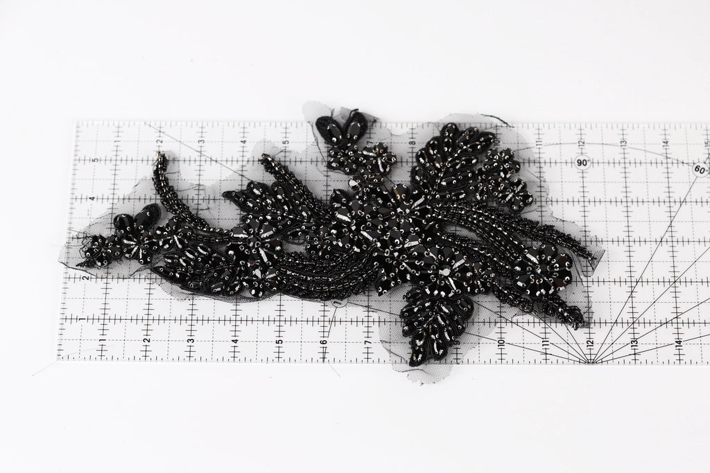 a close-up view of a black, intricate piece of embroidery or beading, with a ruler placed on top of it to measure its dimensions.