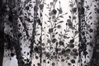 a close-up view of a delicate, intricate black and white lace fabric with a floral pattern. The lace appears to be made of a sheer material, giving it a delicate and elegant appearance. The floral pattern consists of numerous small black flowers and leaves, which are intricately woven into the fabric, creating a visually striking and detailed design.