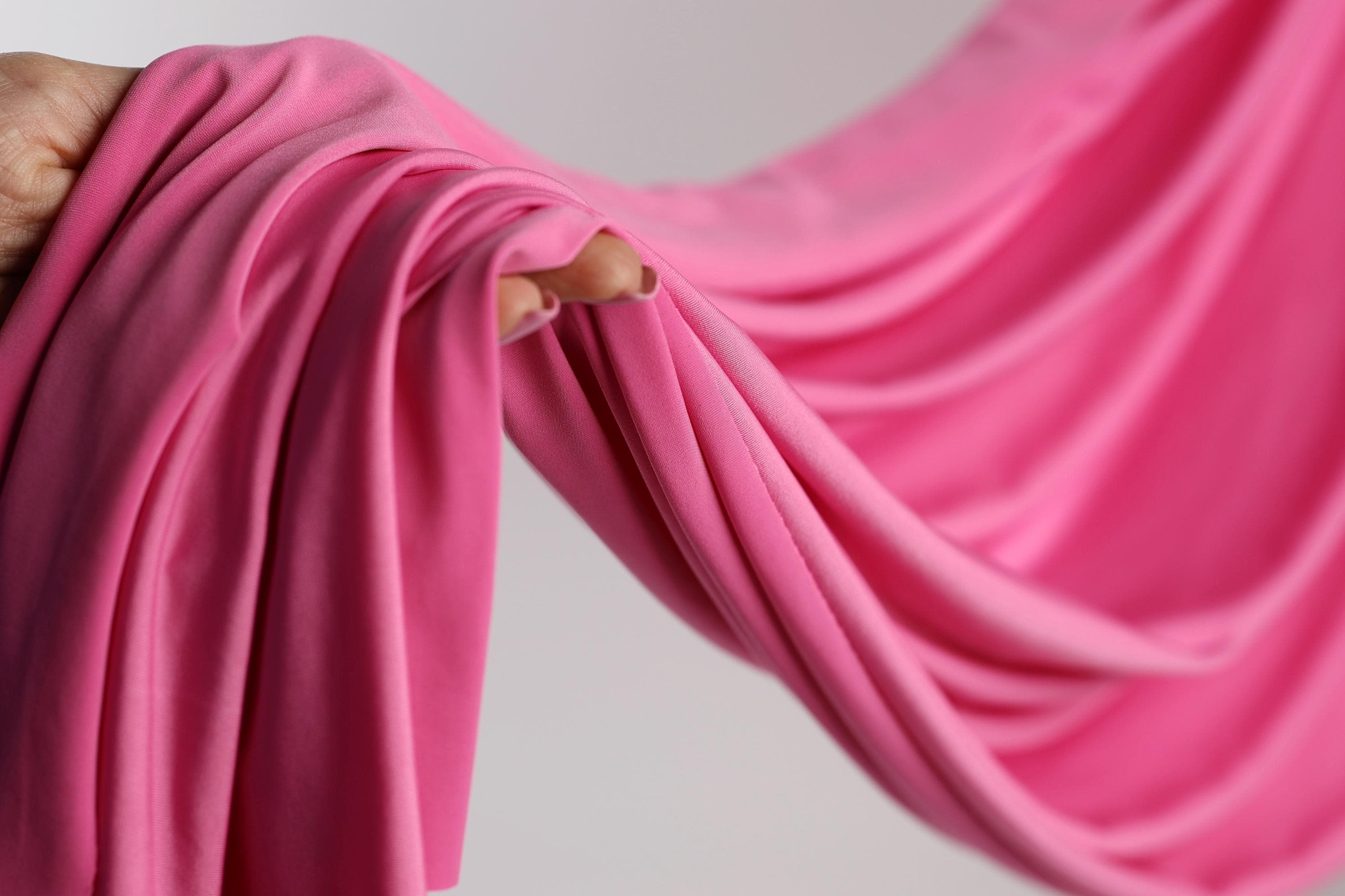 A hand is holding a pink fabric, which appears to be a soft, flowing material. The fabric is being held in a way that it is draped over the hand, creating a sense of movement and softness.