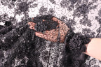 A hand holding a piece of black fabric with a floral pattern, against a background of a white fabric with black floral designs.