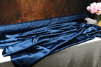 a roll of blue satin fabric on a table, with a vase of pink flowers placed next to it.