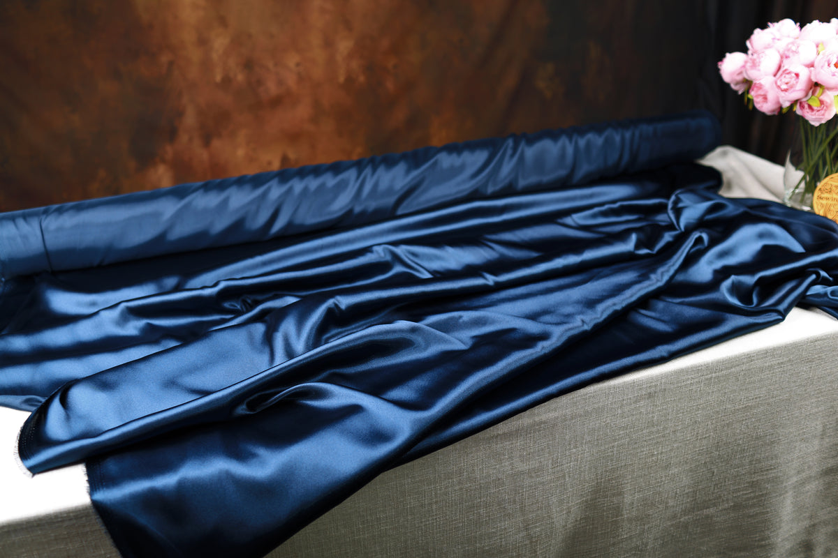 a roll of blue satin fabric on a table, with a vase of pink flowers placed next to it.