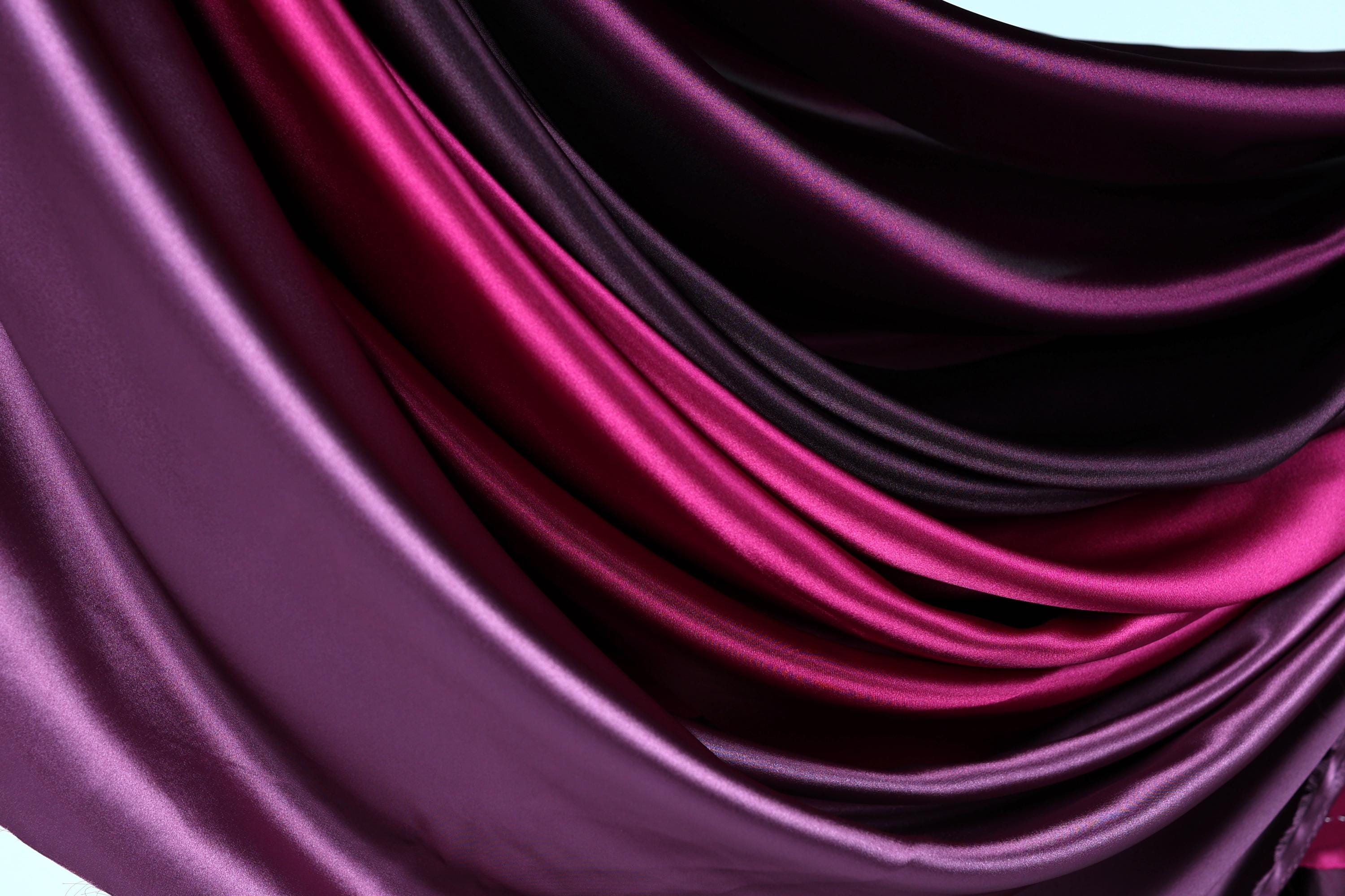 a close-up view of a fabric with a gradient of colors, transitioning from a deep purple to a lighter shade of purple. The fabric appears to be made of satin or a similar smooth, shiny material, and it is folded or draped in a way that creates a sense of depth and texture.