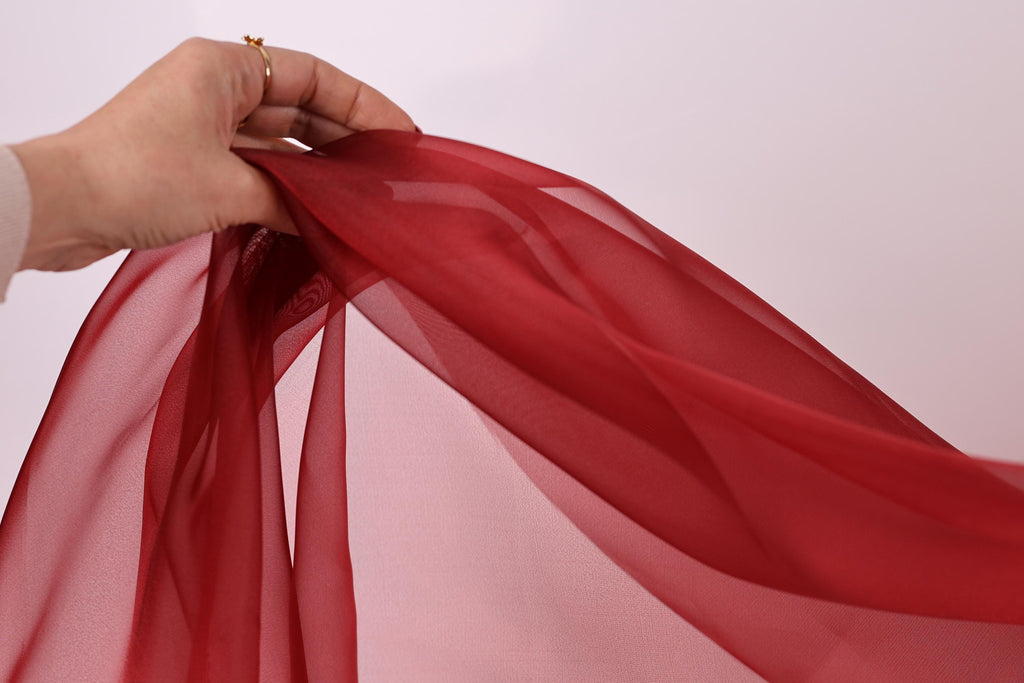 A hand is holding a red fabric, possibly silk or chiffon, against a white background.