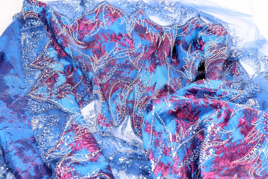 a close-up view of a fabric with a vibrant, colorful pattern of blue, red, and silver sequins or beads. The fabric appears to be a type of lace or sheer material, with a textured and intricate design.