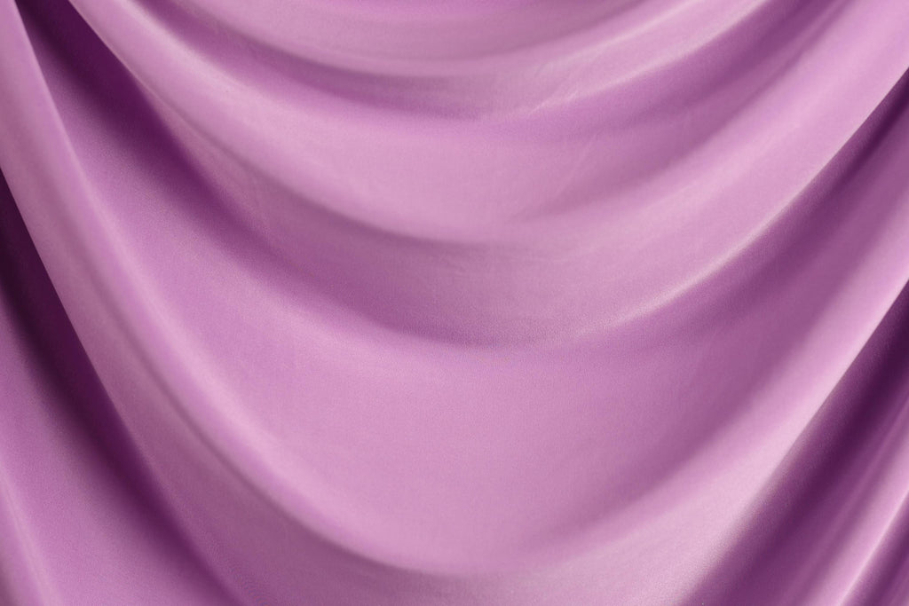 The image is a close-up of a soft, flowing purple fabric, possibly a curtain or garment. The fabric has a smooth, shiny texture and is draped in a way that creates a sense of movement and depth.