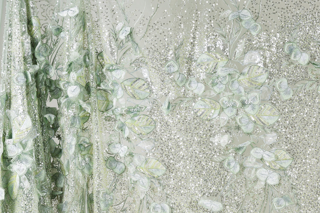 a close-up view of a fabric with a floral pattern, including leaves and flowers in shades of green and white. The fabric appears to be made of a shiny, glittery material, giving it a sparkling and delicate appearance.