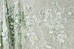 a close-up view of a fabric with a floral pattern, including leaves and flowers in shades of green and white. The fabric appears to be made of a shiny, glittery material, giving it a sparkling and delicate appearance.