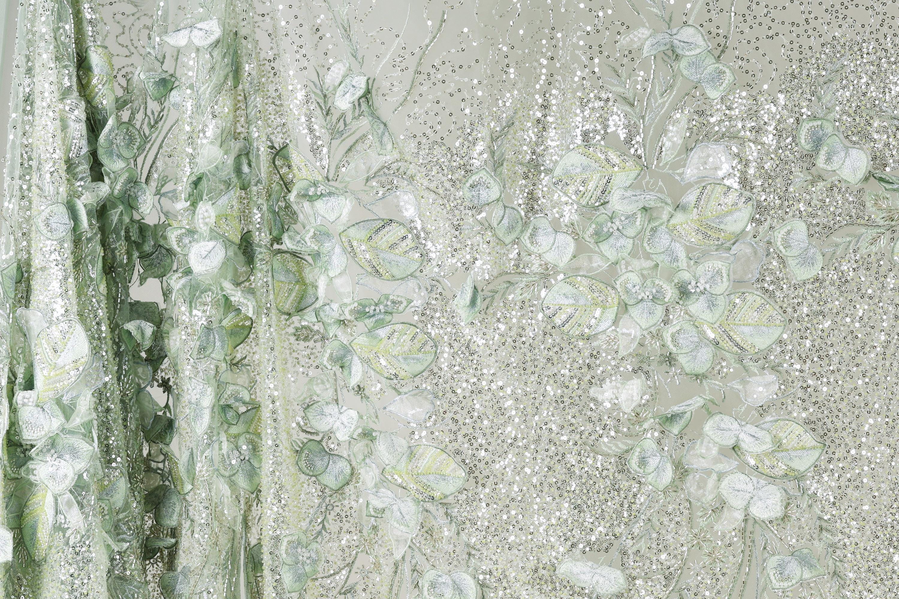 a close-up view of a fabric with a floral pattern, including leaves and flowers in shades of green and white. The fabric appears to be made of a shiny, glittery material, giving it a sparkling and delicate appearance.