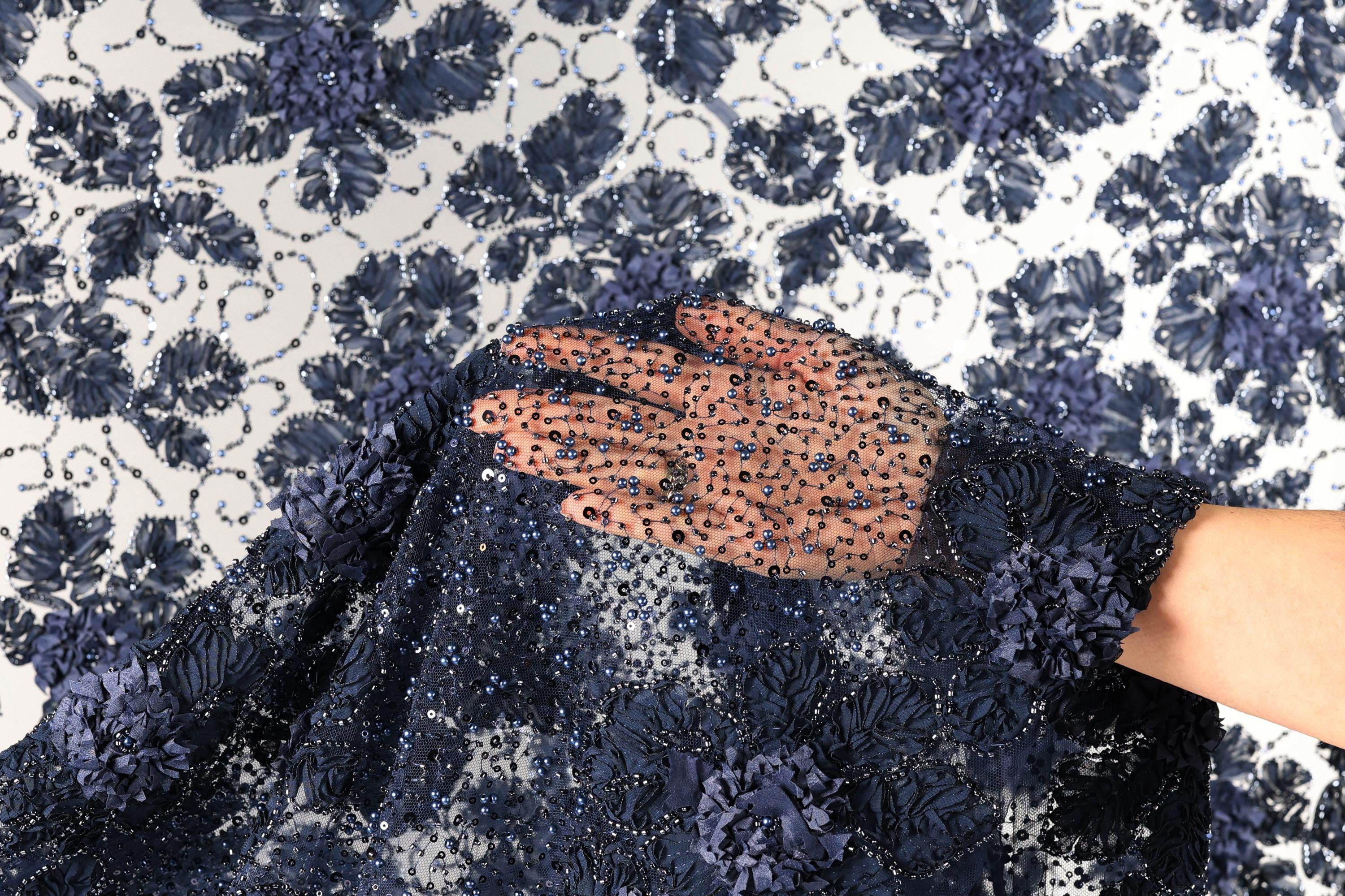 A hand is holding a piece of fabric with a floral pattern and sequins, against a background of a white and blue floral pattern.