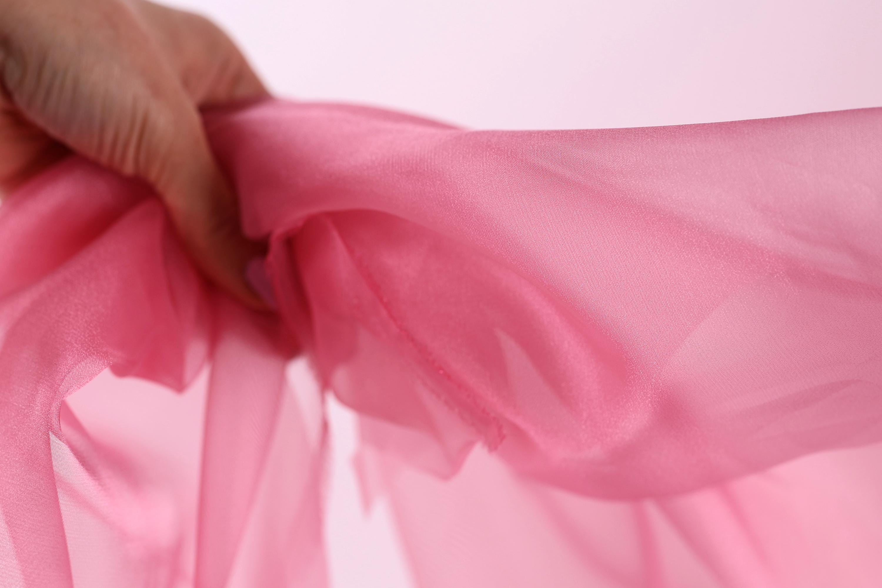 A hand is holding a pink fabric, which appears to be a soft, flowing material.