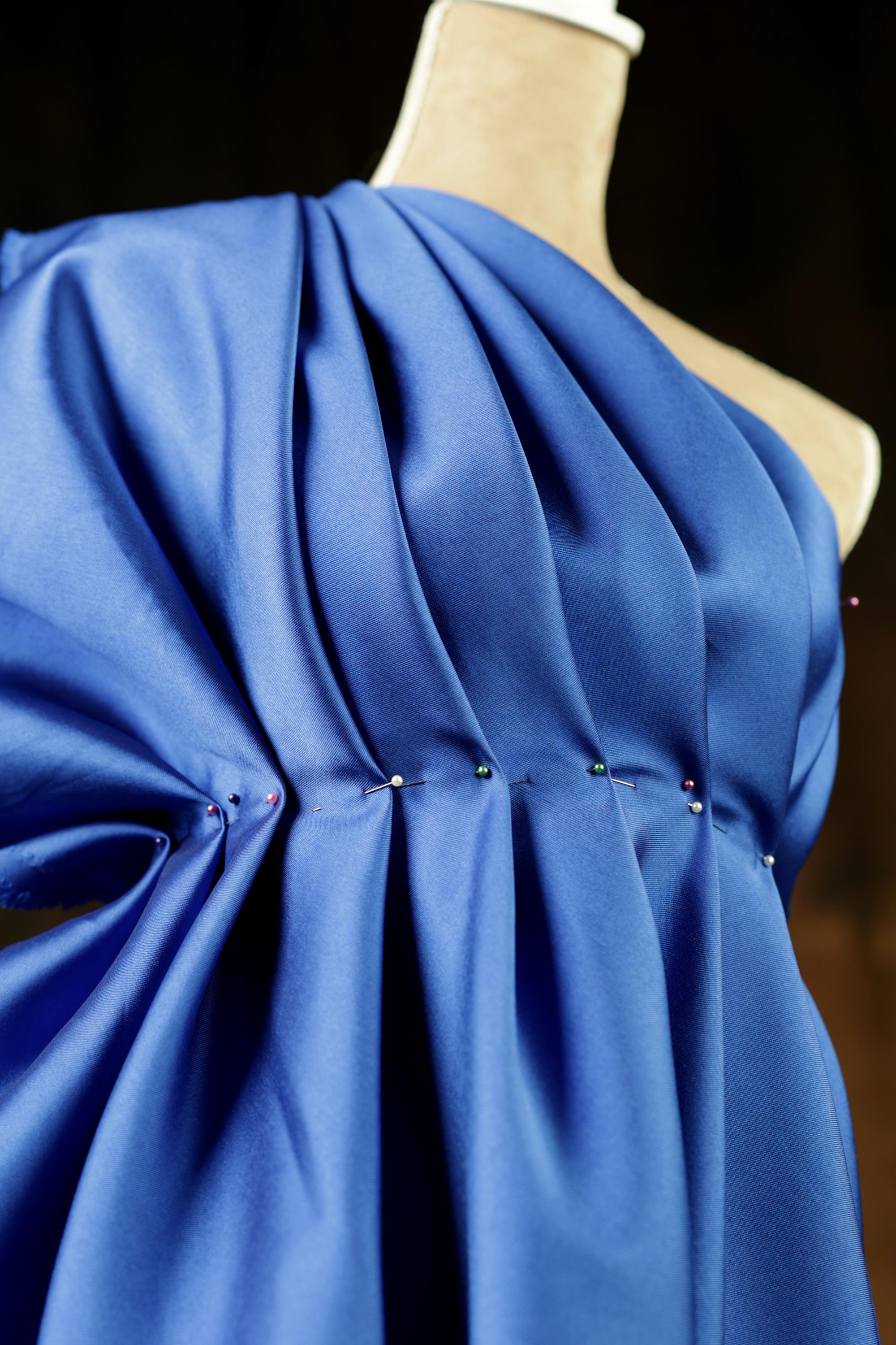 A mannequin wearing a blue satin dress with a ruched neckline and pinned embellishments.