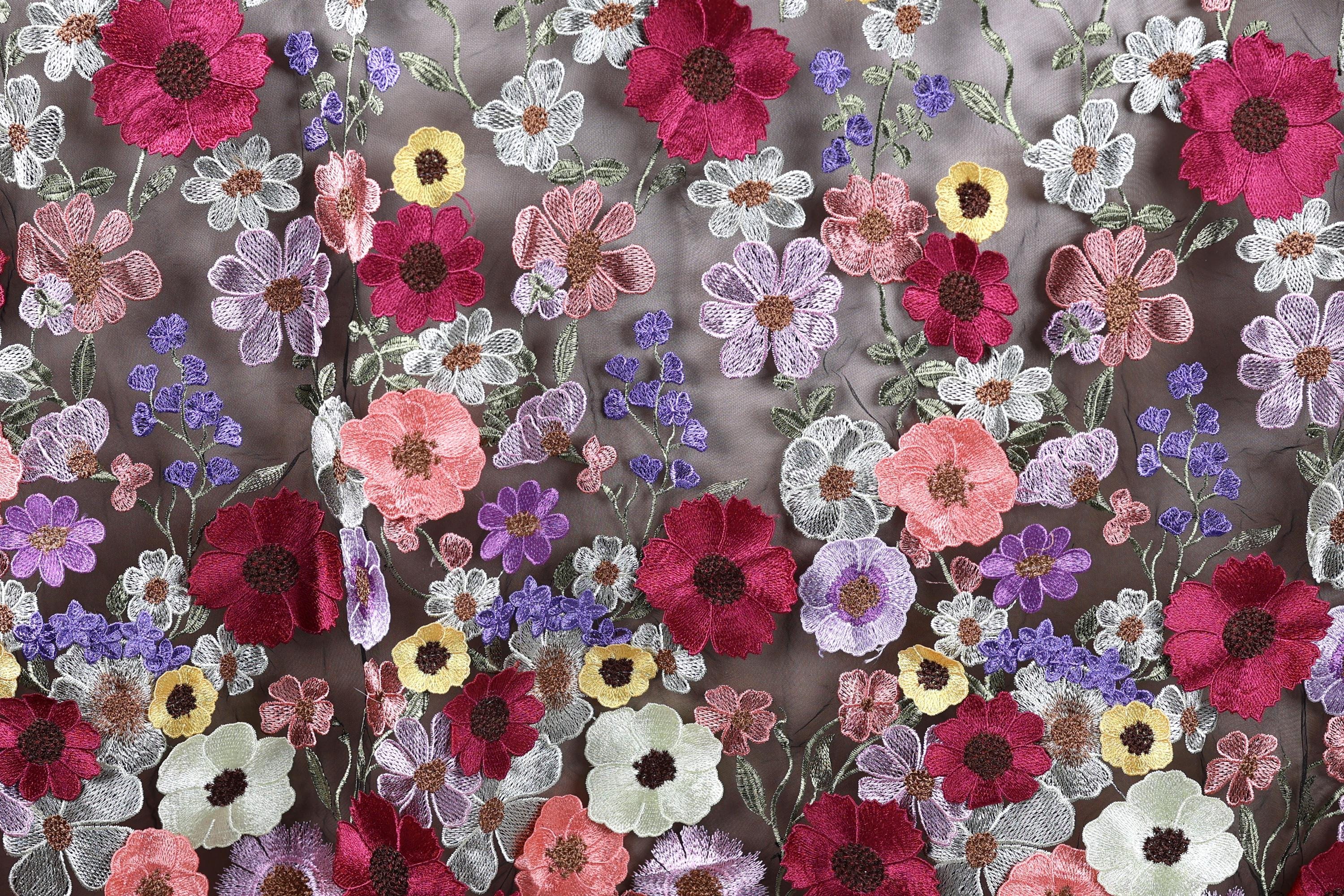 a close-up view of a fabric with a vibrant floral pattern. The fabric is adorned with a variety of colorful flowers, including red, pink, purple, and yellow flowers, as well as green leaves. The flowers are intricately embroidered or stitched onto the fabric, creating a visually striking and detailed design. The image does not contain any text or other discernible objects, and the focus is solely on the floral pattern on the fabric.