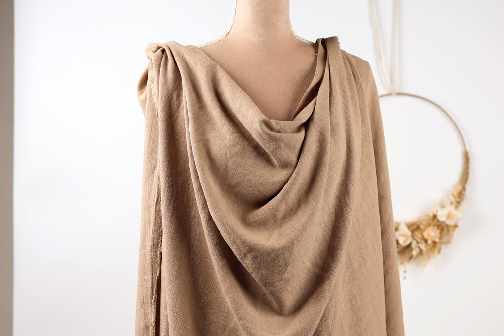 A mannequin wearing a beige draped garment, with a wreath hanging in the background.
