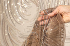 A hand is holding a piece of brown and silver sequin fabric, with the background blurred to focus attention on the fabric.