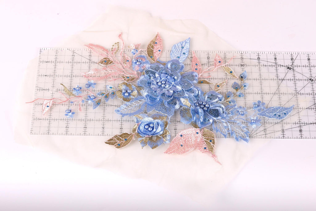 a close-up view of a delicate floral embroidery on a white background. The embroidery is composed of various blue and pink flowers, leaves, and other decorative elements, creating a visually appealing and intricate design.