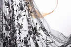 a close-up view of a black, sheer fabric with a floral pattern. The fabric appears to be made of sheer material, giving it a delicate and flowing appearance. The floral pattern consists of black flowers and leaves, which are intricately embroidered or printed onto the fabric. The fabric is spread out, revealing its full length and the details of the floral design.