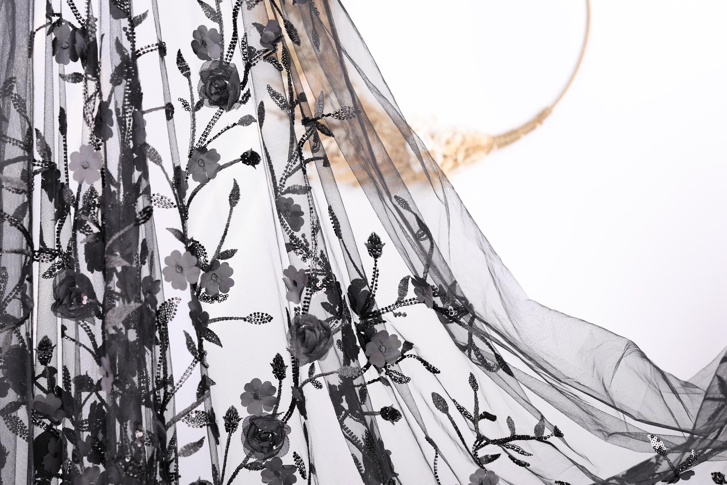 a close-up view of a black, sheer fabric with a floral pattern. The fabric appears to be made of sheer material, giving it a delicate and flowing appearance. The floral pattern consists of black flowers and leaves, which are intricately embroidered or printed onto the fabric. The fabric is spread out, revealing its full length and the details of the floral design.