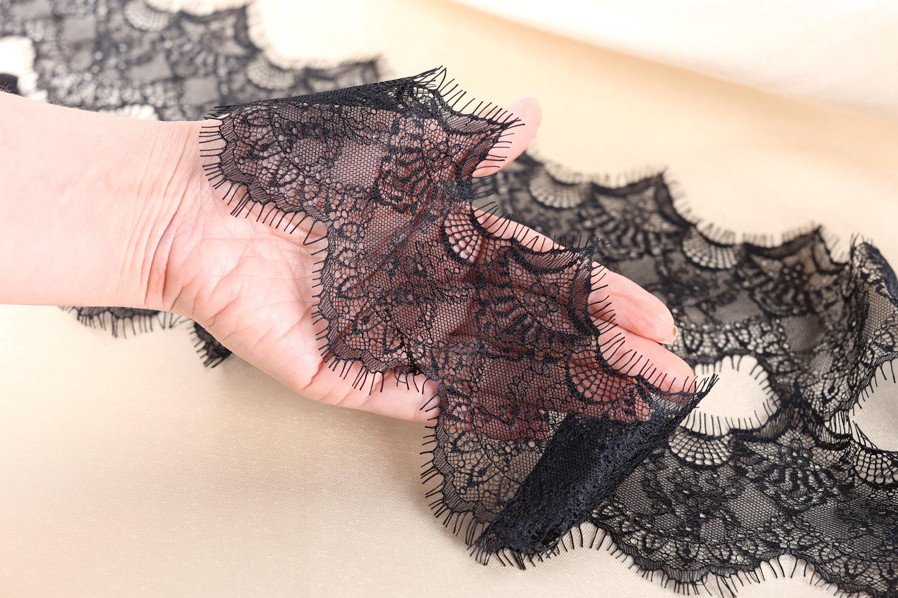 A hand is holding a piece of black lace with a floral pattern, which appears to be a decorative fabric.