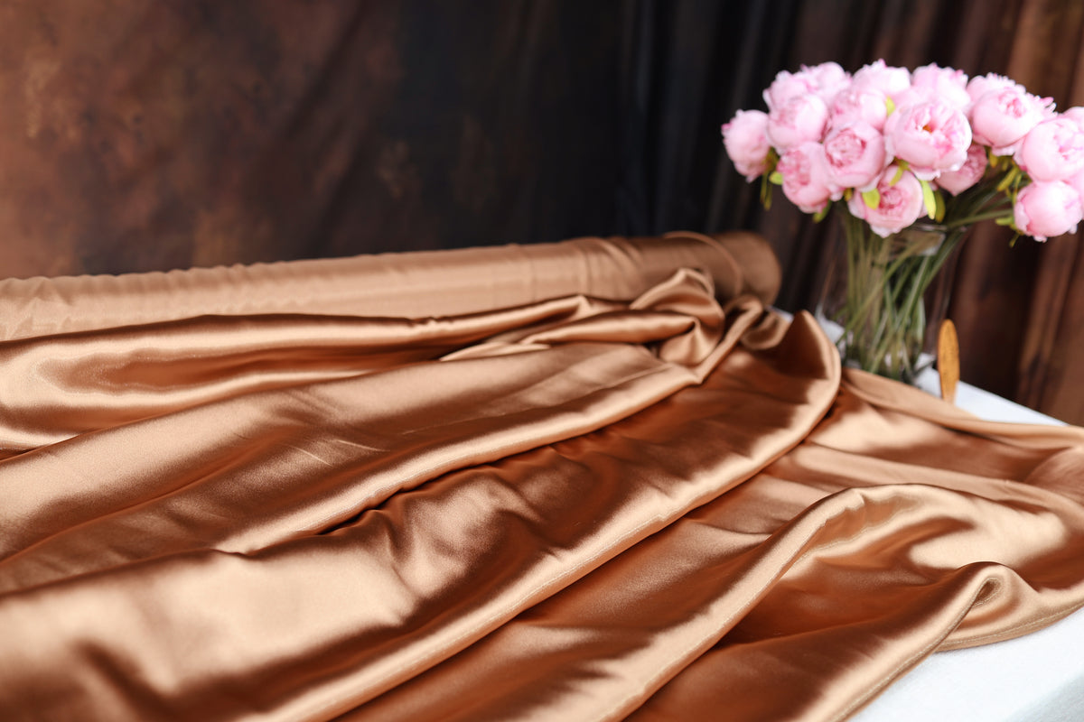 a close-up of a piece of shiny, golden fabric, possibly silk or satin, with a vase of pink flowers placed next to it on a table.