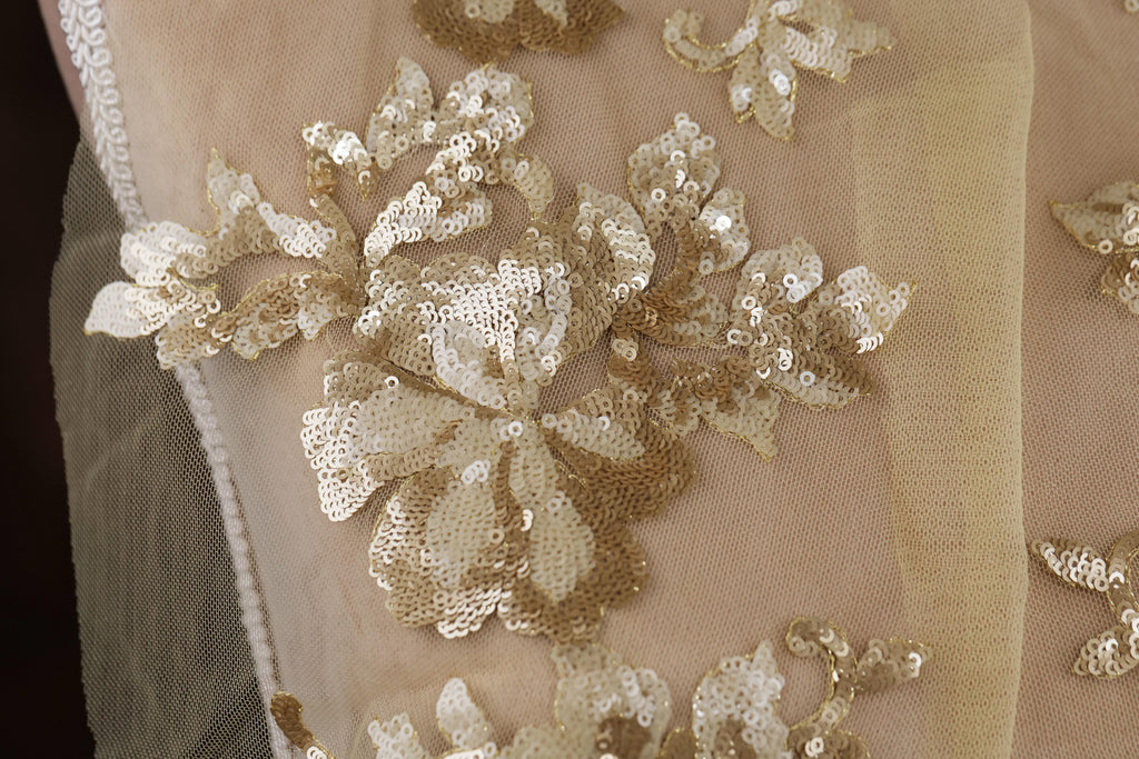 a close-up view of a delicate, intricate piece of fabric adorned with a floral pattern made of small, shiny gold sequins. The fabric appears to be a light-colored material, possibly lace or sheer, with a delicate, elegant design.
