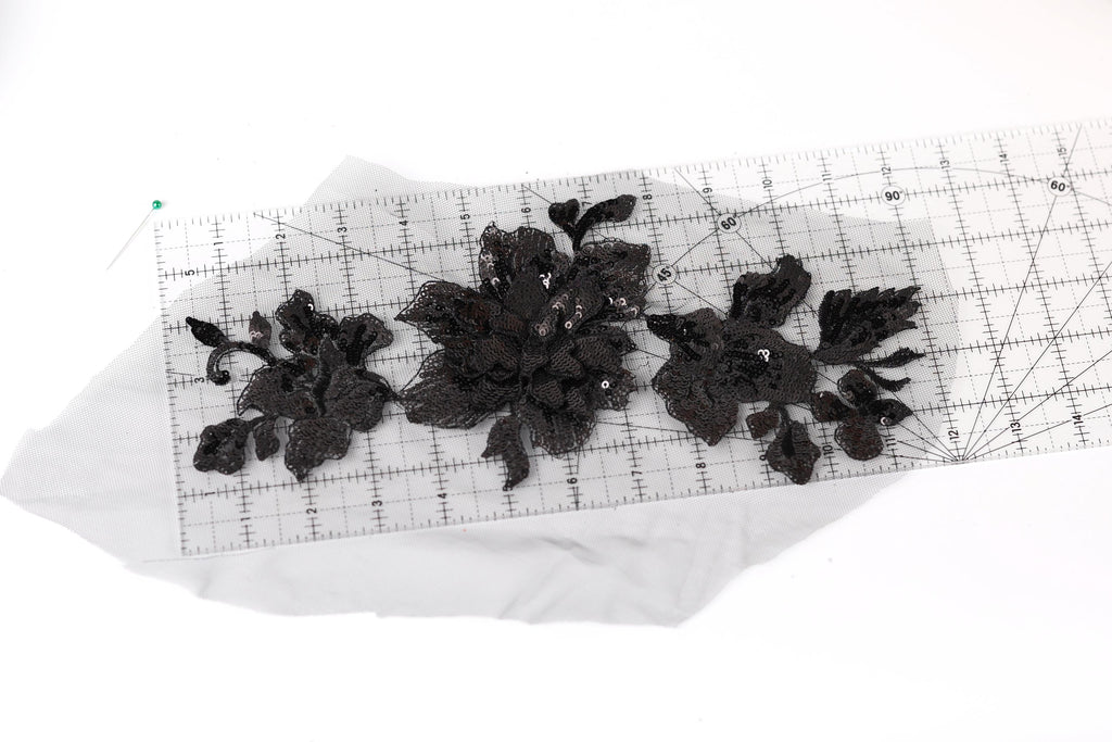 a close-up view of a piece of fabric with black lace or embroidery designs, placed on a transparent ruler or measuring mat.